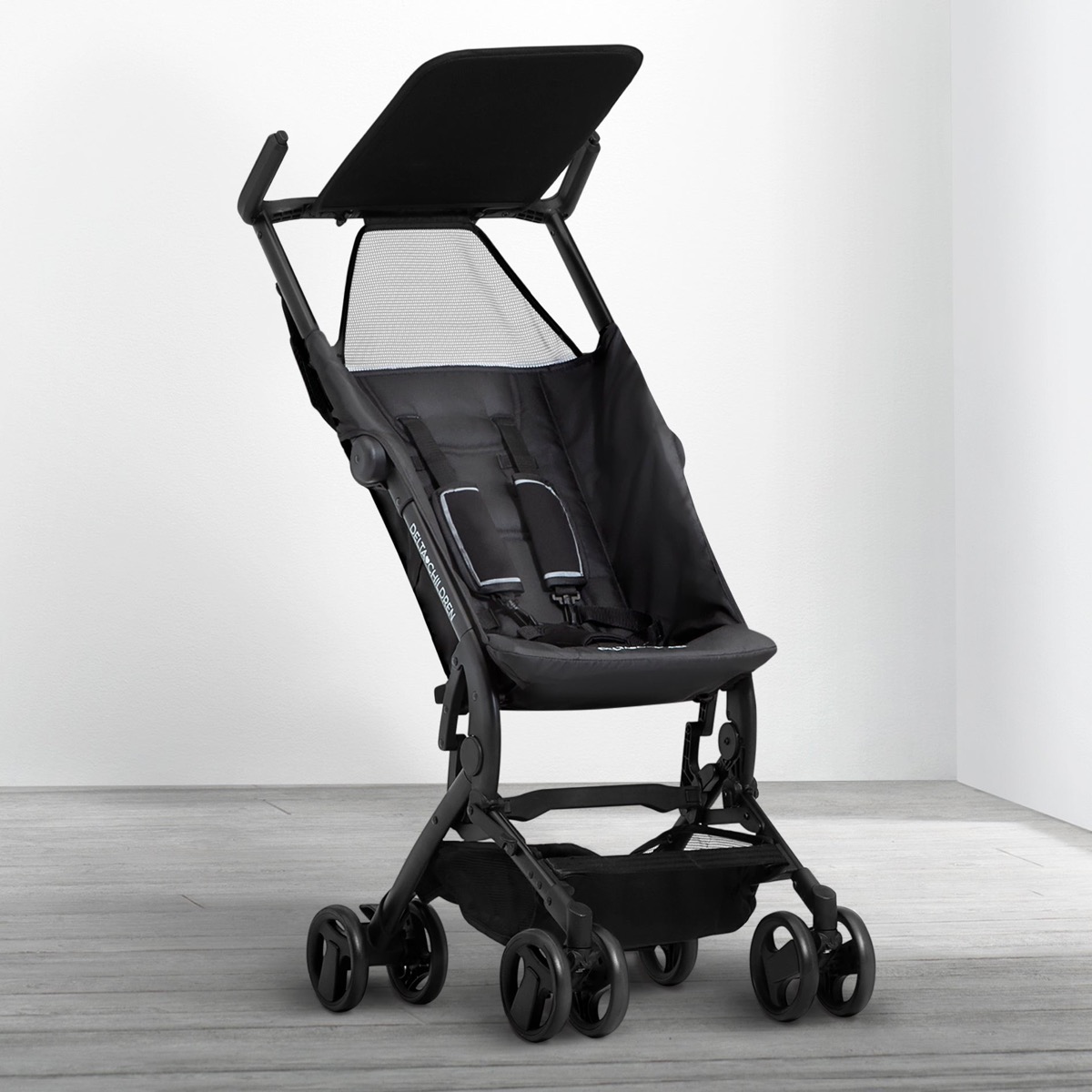 The Clutch stroller in Black standing open in a minimalist room corner, side view showing the slim frame, harness pads, and storage basket