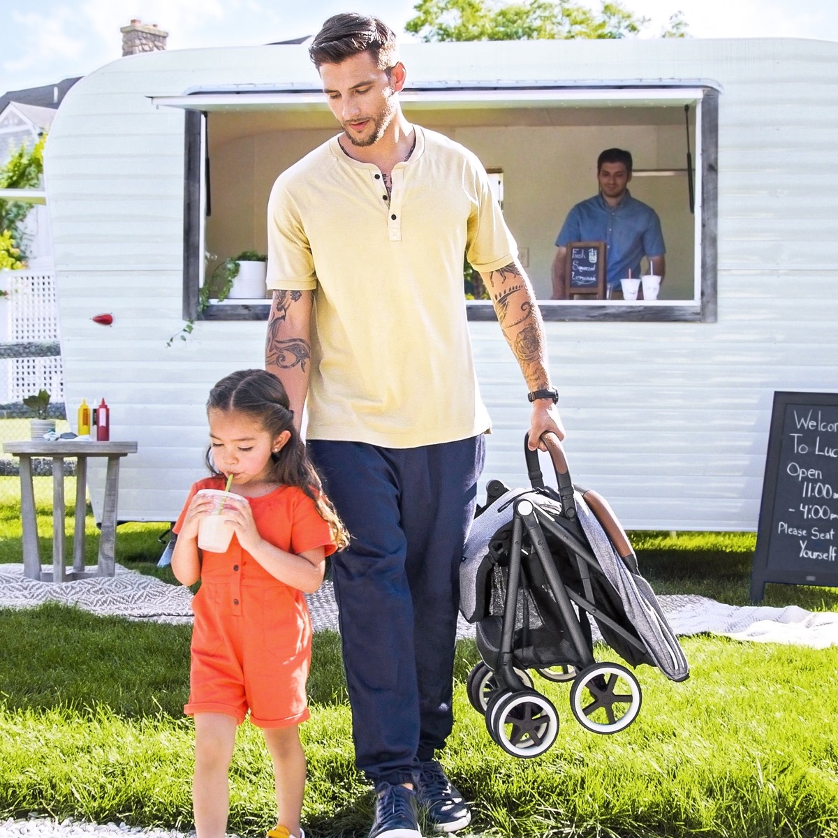 An adult carrying the folded gray Evenflo Gold Otto stroller one-handed by its bumper bar while walking outdoors with a toddler