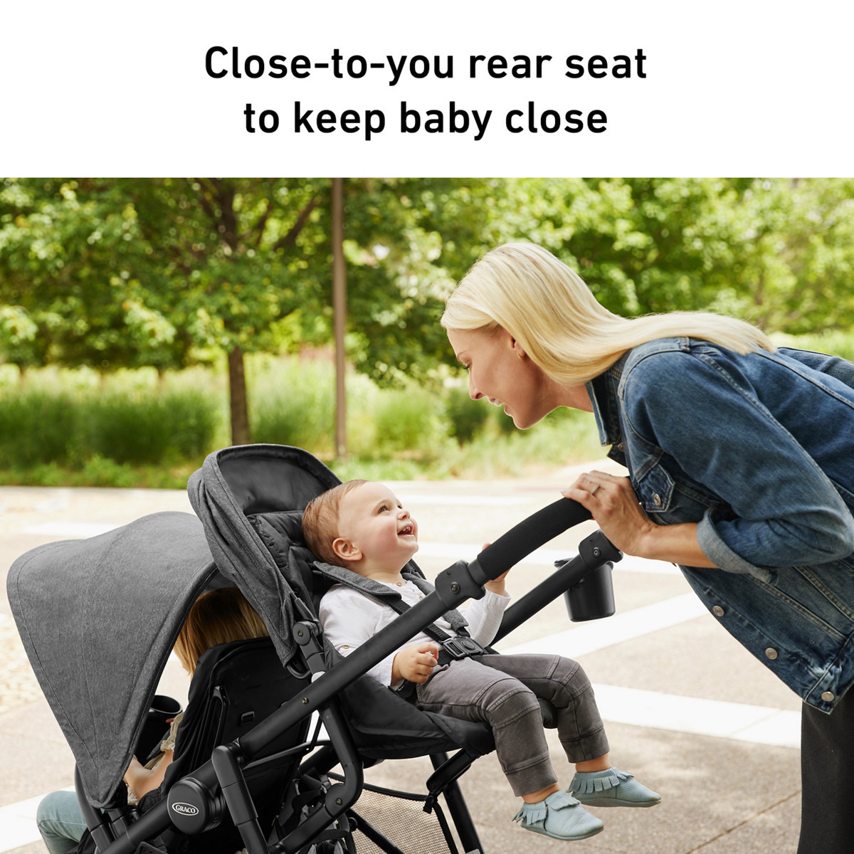 A smiling woman leans over the handlebar to look at a laughing baby in the elevated close-to-you rear seat while an older child sits in the front seat, on a tree-lined walkway, with the burned-in headline 'Close-to-you rear seat to keep baby close'