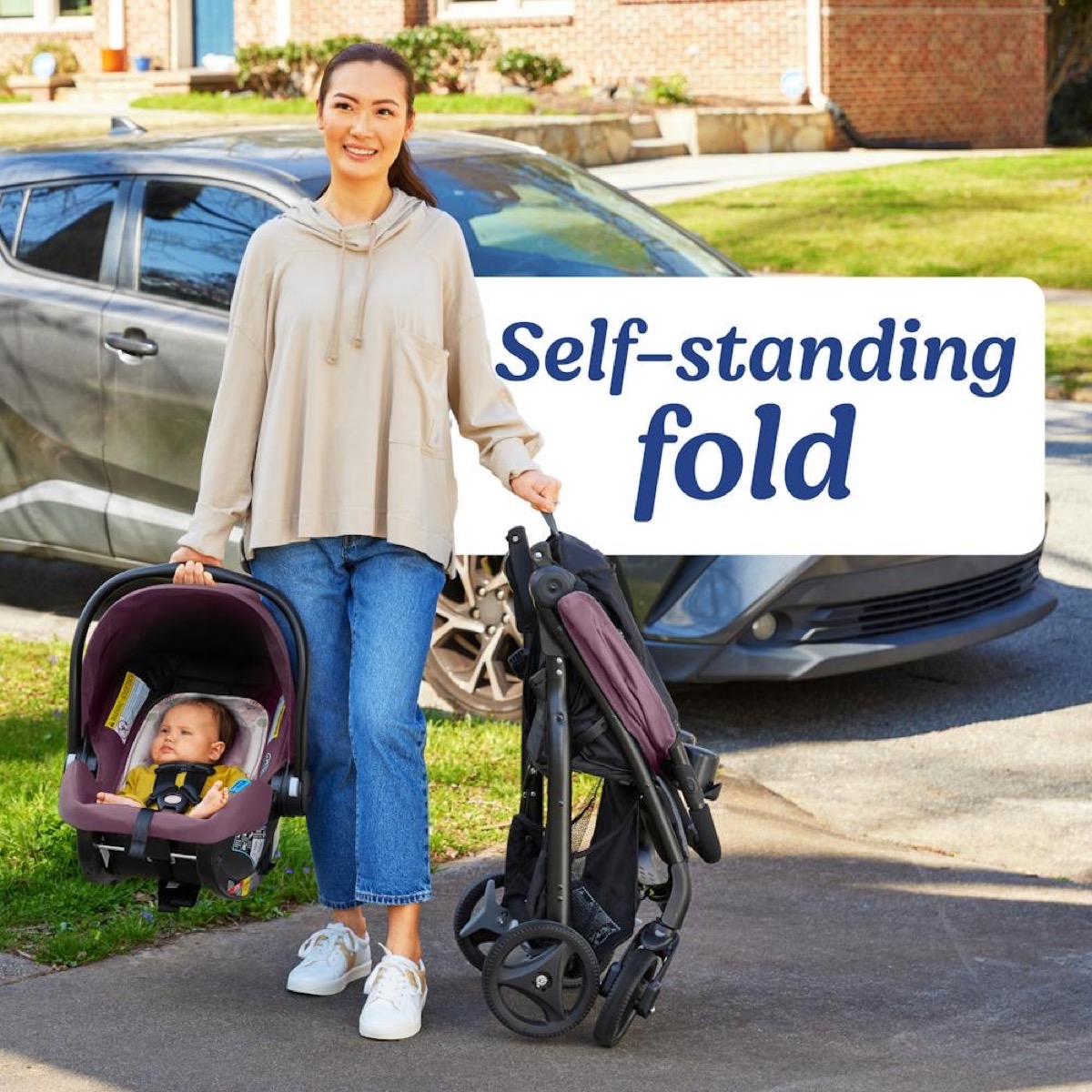 Outdoor photo: a caregiver holds the folded Verb stroller upright by its carry strap (self-standing fold, wheels down, plum Merrick fabric visible) while carrying the matching SnugRide infant car seat with a baby at her side, in front of a parked car; overlaid text 'Self-standing fold'