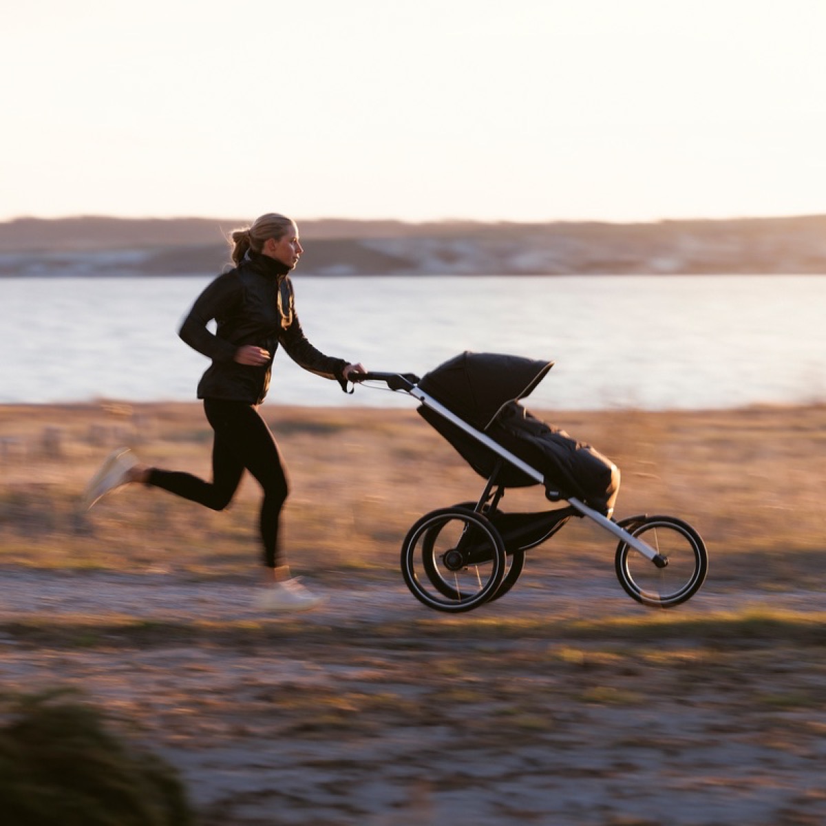 A woman jogging along a waterfront path at sunset while pushing a Thule Glide 3 stroller