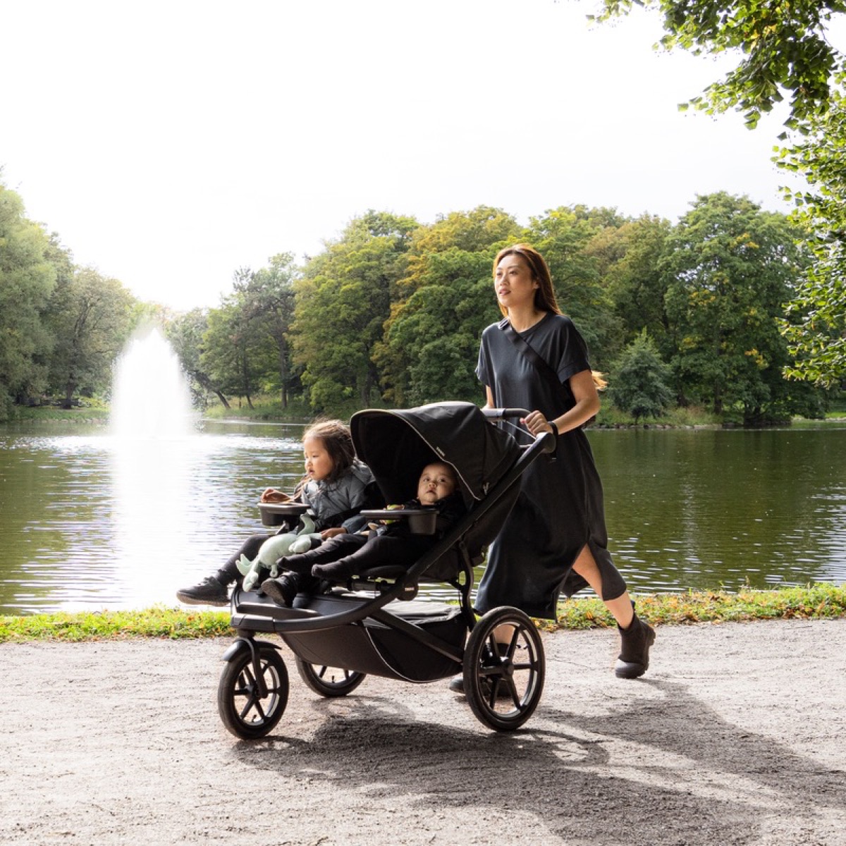 A caregiver walking through a park with two children seated side by side in the black Thule Urban Glide 3 double stroller